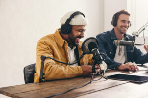 Two college podcasters laughing and having a good time in a studio. Two happy young men co-hosting a live audio broadcast. Two male content creators recording an internet podcast.