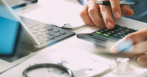 Laptop and stethoscope on desk with hands using calculator